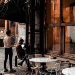 Historic Districts - Facade of cafe with large windows and tables on sidewalk along road and faceless people resting in city